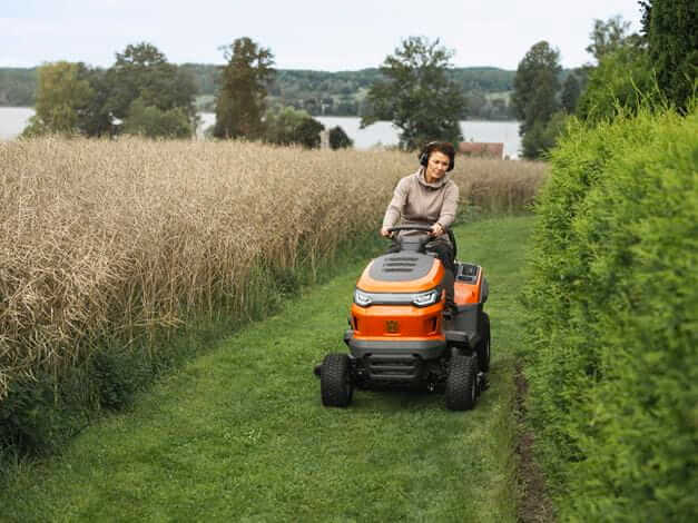 Traktor TS 215T Husqvarna w kolorze pomarańczowym, z czarnymi detalami, prowadzi osoba na trawiastym torze. Ujęcie z boku ukazuje kółka i prostą konstrukcję. Tło stanowi pole i zarośla, co wskazuje na użytkowanie w ogrodzie.