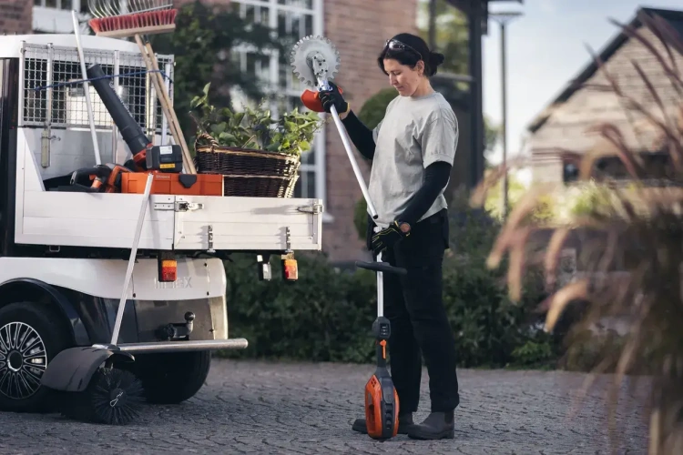 Kobieta w rękawicach używa podkaszarki Husqvarna 525iLK z metalową tarczą i ergonomiczną rączką. W tle ogrodowy pojazd z narzędziami i roślinami w koszu, na tle zieleni.