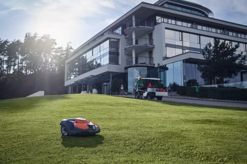 Niebo-pomarańczowy robot koszący Automower® 520 Epos Husqvarna na trawie przed nowoczesnym budynkiem. W tle osoby używające kosiarek ręcznych. Urządzenie pracuje na lekko nachylonym terenie.