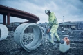 Mężczyzna w żółtej kurtce i hełmie używa pionowej zagęszczarki Husqvarna LT 6005 przy dużej betonowej rurze na placu budowy, w tle materiały i konstrukcje budowlane.