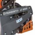Zagęszczarka Husqvarna LT 6005 z widoczną szaro-czarną obudową i pomarańczowymi elementami. Ujęcie frontalne ukazuje napisy z logo oraz oznaczenia funkcyjne na boku urządzenia. Widać metalowe detale mocujące i układ roboczy.
