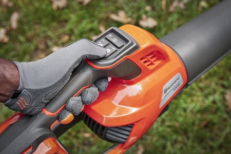 Dmuchawa akumulatorowa Husqvarna 230iB, widok zbliżony na uchwyt z przyciskami sterującymi. Uchwyt w czarnym kolorze z elementami pomarańczowymi, w dłoni użytkownika w rękawicach. Tło w naturalnym otoczeniu, wśród trawy.