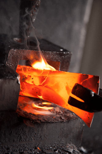Zgrzewany metal w ogniu na kowadle, o intensywnej czerwonej barwie. Widać dym unoszący się z gorącej powierzchni, kształt metalu jest nieregularny, a obok widoczny uchwyt narzędzia. Ujęcie zbliżone, z różnymi materiałami w tle.
