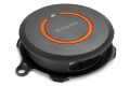Husqvarna Automower FlexiFence, okrągły element w kolorze czarnym z pomarańczowym wykończeniem na górze. Widok z góry, pokazujący logo Husqvarna oraz przycisk, z wyraźnym zdobieniem. Podstawa przystosowana do montażu.
