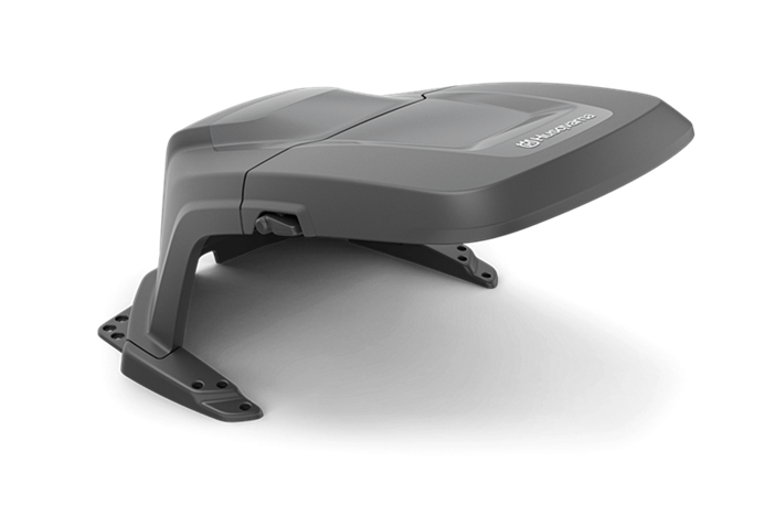 Domek - osłona dla Automower serii Nera Husqvarna w kolorze szarym, stylizowany z wyraźnymi krawędziami. Złożony, o aerodynamicznej formie, ma logo Husqvarna. Widok z boku, na jednolitym tle, umożliwia pełne zaprezentowanie detali konstrukcji.