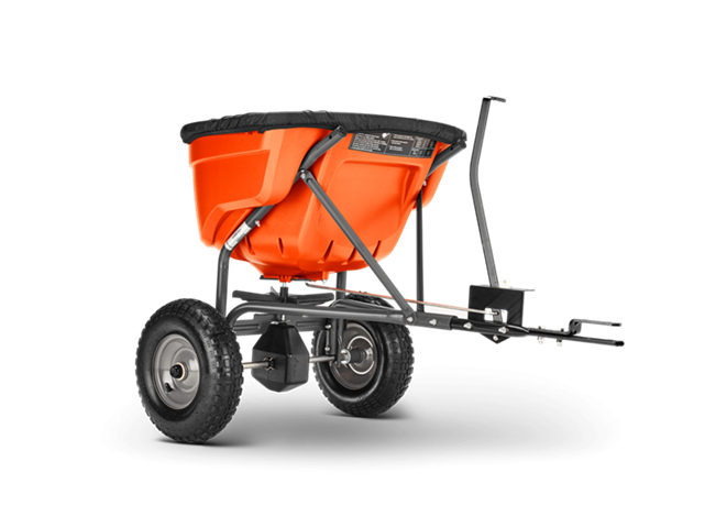 Rozsiewacz Husqvarna 75 kg w kolorze pomarańczowym, umieszczony na metalowej ramie z dwoma dużymi kołami. Ujęcie z boku, pokazujące pojemnik z czarną osłoną i mechanizm do montażu. Produkt przeznaczony do traktora lub ridera.