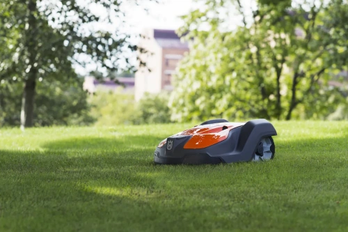 Robot koszący automatyczny Husqvarna Automower® 550 w kolorze pomarańczowym i szarym, leżący na trawie. Ujęcie z boku, pokazujące zaokrąglony kształt i charakterystyczne logo. Tło z drzewami i budynkiem w oddali, w naturalnym otoczeniu.