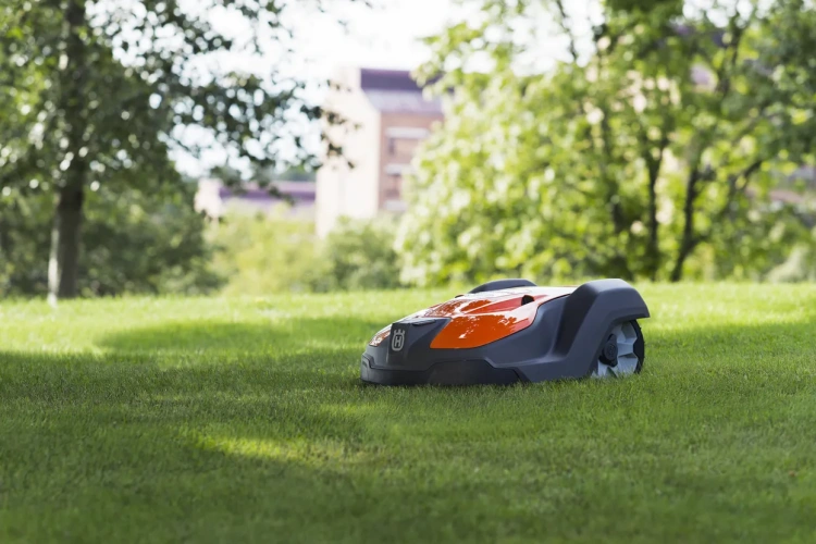 Robot koszący automatyczny Husqvarna Automower® 550 w kolorze pomarańczowym i szarym, leżący na trawie. Ujęcie z boku, pokazujące zaokrąglony kształt i charakterystyczne logo. Tło z drzewami i budynkiem w oddali, w naturalnym otoczeniu.
