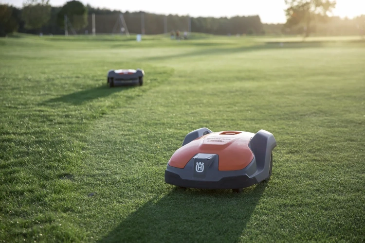 Robot koszący Husqvarna Automower® 550 w kolorze szarym z pomarańczowymi akcentami, widoczny w zbliżeniu na trawie. Ujęcie z boku, produkt porusza się po gładkiej, dobrze przystrzyżonej powierzchni. Tło przedstawia pole, otoczone drzewami.