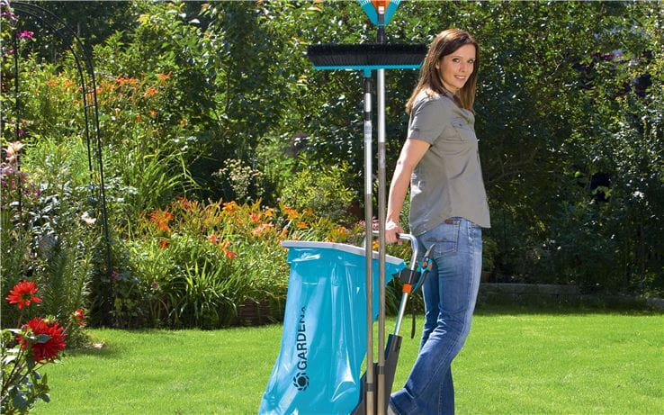 Wózek ogrodowy GARDENA w kolorze niebieskim, z otwartym koszem, umiejscowiony na trawniku. Produkt trzymany jest przez kobietę w szarej koszulce, na tle kolorowego, kwiatowego ogrodu. Widoczna miotła i akcesoria ogrodowe.