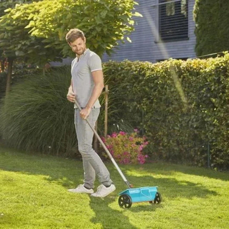Siewnik Gardena 3 l w kolorze błękitnym z czarnymi kółkami, używany przez mężczyznę w jasnej koszulce na trawie w ogrodzie z krzewami i kwiatami w tle.