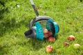 Zbieraczka do owoców Gardena w kolorze czarnym i błękitnym, z siatkowymi ściankami, leży na trawie. Widać kilka jabłek w obrębie zbieraczki oraz na trawie. Ujęcie jest z boku, ukazując detal konstrukcji oraz materiał.