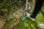 Szpadel półokrągły Gardena w jasnoniebieskim kolorze z metalowym uchwytem, włożony w zieleń trawnika, z nogą użytkownika opartą na uchwycie. Roślinność w tle dodaje naturalnego kontekstu. Produkt w użyciu, ukazujący jego funkcjonalność.