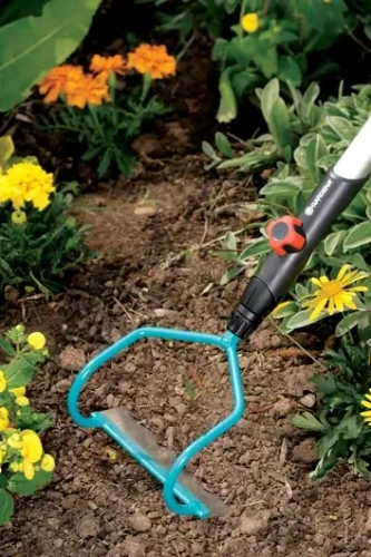 Motyczka Gardena Combisystem 16 cm w błękicie z ergonomicznym uchwytem i stalowym ostrzem do precyzyjnej pielęgnacji gleby, leży w ziemi otoczona żółtymi i zielonymi kwiatami.