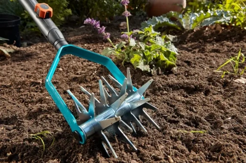 Kultywator gwiazdowy Gardena o średnicy 14 cm, z metalową głowicą i niebieską rączką z tworzywa sztucznego. Produkt ułożony na ziemi w ogrodzie, z widocznymi roślinami w tle. Zbliżenie na detale ostrzy kultywatora.