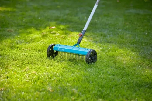 Wertykulator do trawy na kółkach Gardena, o szerokości 32 cm, w kolorze błękitnym i czarnym, z metalowymi ostrzami, stoi na trawie. Możliwe widoczne logo. Produkt pokazany jest w użyciu, na równym, zielonym tle.
