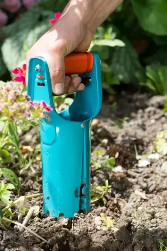 Sadzarka do cebul kwiatowych Gardena w błękitnym kolorze z pomarańczowym uchwytem, trzymana w ręce. Produkt jest włożony w ziemię, a w tle widoczne są kwiaty i zieleń. Uchwyt ergonomiczny z otworami ułatwia trzymanie i obsługę.