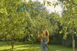 Teleskopowy trzonek Gardena 160-290 cm z niebieską końcówką, używany przez kobietę sięgającą w górę w zielonym ogrodzie pełnym drzew i trawy.