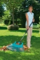 Trzonek Aluminiowy Gardena w kolorze błękitnym, stojący w dłoniach użytkownika na trawniku. Na końcu trzonka znajduje się zgarniacz do liści. Otoczenie to zielony ogród z drzewami i krzewami w tle, na którym leży stos liści.
