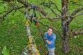 Nożyce Gardena z teleskopowym trzonkiem do cięcia gałęzi, szaro-pomarańczowe. Mężczyzna używa ich na tle zielonej trawy i drzew, widok z góry na narzędzie i gałąź.