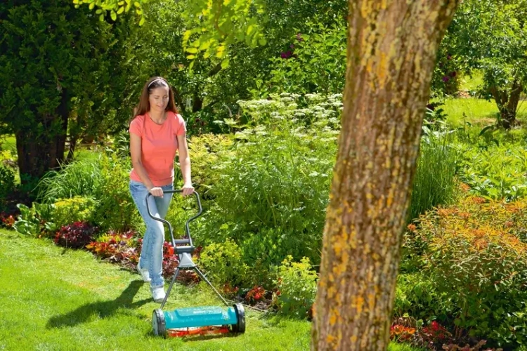 Kosiarka bębnowa Gardena Classic 400 w użyciu. Użytkowniczka w różowej koszulce kosi trawę w ogrodzie z zielenią i kwiatami. Kosiarka zielona z metalowymi detalami, widok z boku.