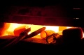 Wnętrze pieca z żeliwnymi elementami i pomarańczowymi płomieniami. Metalowe szczypce leżą obok gorących przedmiotów na ciemnym tle podkreślającym światło ognia.