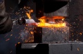 W powiększeniu widoczny proces obróbki metalu, gdzie pręt jest formowany w gorącym piecu kowalnym. W tle delikatne iskry i płomienie. Stalowy pręt trzymany jest w rękawicy, z bliska ukazując intensywny żar procesu.