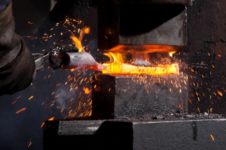 W powiększeniu widoczny proces obróbki metalu, gdzie pręt jest formowany w gorącym piecu kowalnym. W tle delikatne iskry i płomienie. Stalowy pręt trzymany jest w rękawicy, z bliska ukazując intensywny żar procesu.