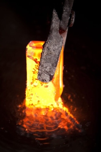 Zdjęcie przedstawia gorący kawałek metalu w pomarańczowym kolorze, który jest podtrzymywany przez narzędzie o metalowym uchwycie. Tło jest ciemne, co kontrastuje z intensywnym odcieniem metalu. Widoczna jest faktura powierzchni metalu.