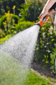 Sitko do zraszaczy Gardena w użyciu, trzymane w dłoni. Z bliska widać strumień wody rozpryskujący się w ogrodzie. Rączka w kolorze pomarańczowym wykonana z tworzywa. Tło z zielenią roślinności, słoneczne warunki.