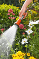Sitko do zraszaczy Gardena w kolorze pomarańczowym i szarym, uchwycone z boku w ręce użytkownika. Produkt używany w ogrodzie, z wodą wypływającą z dyszy, otoczenie pełne kwiatów i zieleni. Widoczne detale uchwytu i strumienia wody.