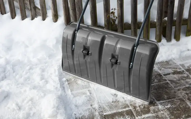 ClassicLine szufla do śniegu Gardena w kolorze szarym, stojąca na podłożu pokrytym śniegiem. Widoczne uchwyty do trzymania oraz plastikowa konstrukcja. Ujęcie z przodu, ukazujące detal krawędzi roboczej. Tło: drewniane ogrodzenie.