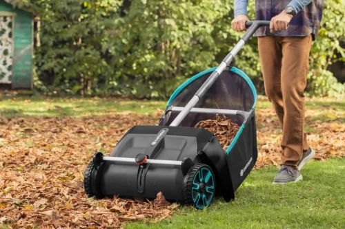 Zbieracz liści i trawy Gardena, czarny z niebieskimi detalami, pełen liści, stojący na trawie. Użytkownik trzyma uchwyt. Duże koła ułatwiają manewrowanie. Tło to zielona roślinność.