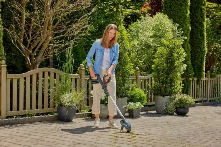 Akumulatorowa szczotka Gardena z ergonomicznym uchwytem i niebieskim kolorem, używana przez kobietę do czyszczenia fug na kamiennej powierzchni w otoczeniu zieleni.