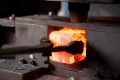 Rozgrzany metal umieszczony w prasie do kucia, trzymany narzędziem przez operatora. Tło ciemne, metalowe z resztkami materiału, podkreślające pomarańczowe światło gorącego metalu.