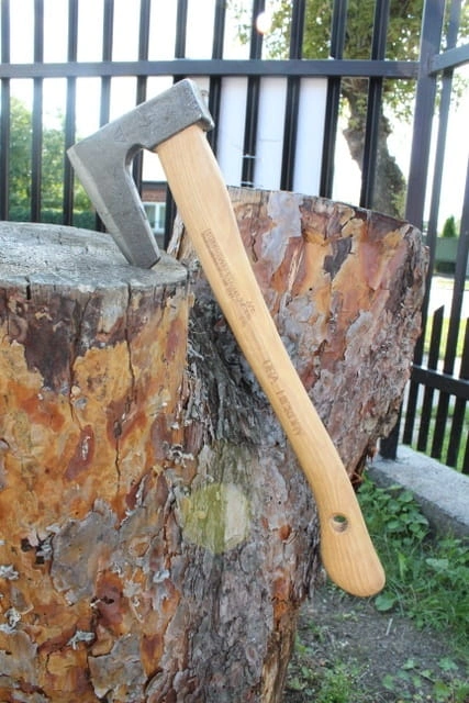 Toporek myśliwski Krumpholz 0.3kg z metalową głowicą i drewnianą rączką o jasnym wykończeniu, leżący na pniu drzewa. Widoczny drobny detal logo na głowicy. Tło naturalne, zielone, z ogrodzeniem w oddali.