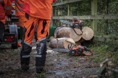 Pomarańczowa pilarka akumulatorowa Husqvarna 540i XPG BT z metalowym łańcuchem stoi obok pni drzew na tle lasu, z widocznym logo marki oraz intensywnie kolorowymi roboczymi ubraniami.