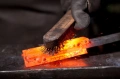 Wizja bliska na gorący, pomarańczowy kawałek metalu, który jest szczotkowany czarną szczotką. Metal leży na stalowej powierzchni, a w tle widać rozmyte ciemne tło. Ręka w czarnej rękawicy wpływa na teksturę i wygląd metalu.