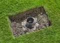 Regulator ciśnienia umieszczony w wykopie z gruntu, otoczony drobnymi kamieniami. Widok z góry, prezentujący jego okrągły kształt oraz dwa wystające węże. Tło stanowi zielona trawa wokół wykopu.