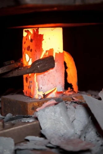 W piecu hutniczym znajduje się rozgrzewany metal w formie słupa, który świeci intensywnym pomarańczowym światłem. Widoczne są fragmenty odpryskującego materiału w otoczeniu, a podstawa jest zwrócona w kierunku narzędzia. Tło jest ciemne.