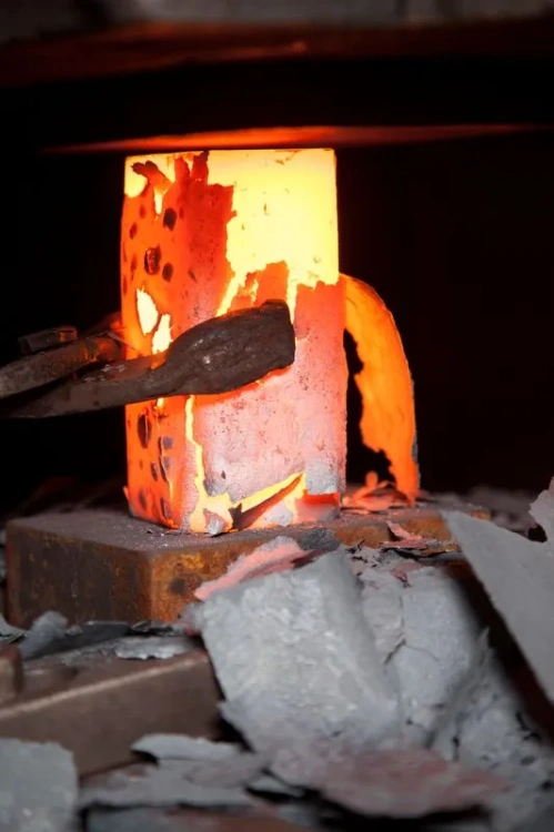 W piecu hutniczym znajduje się rozgrzewany metal w formie słupa, który świeci intensywnym pomarańczowym światłem. Widoczne są fragmenty odpryskującego materiału w otoczeniu, a podstawa jest zwrócona w kierunku narzędzia. Tło jest ciemne.