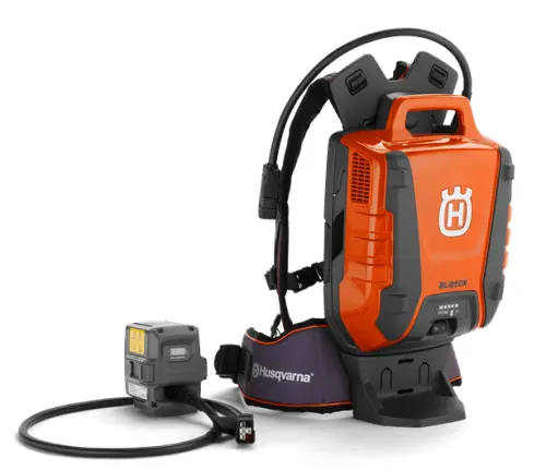 Akumulator plecakowy BLi950X w kolorze pomarańczowym, z metaliczną fakturą. Stojący, z wyraźnym logo Husqvarna. Obok znajduje się jednostka zasilająca z wyświetlaczem. Produkt jest nowy, z widocznymi detalami takimi jak mocowanie i rączka.