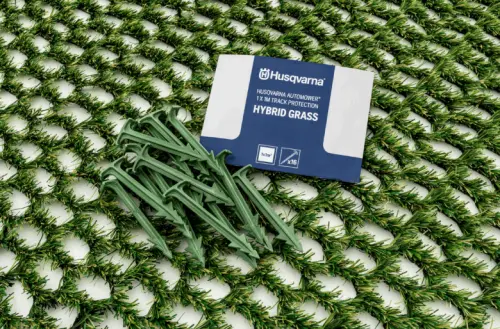Hybrydowa trawa Husqvarna Automower na zielonym tle z bieżnikiem. Widoczne elementy to zielone klamry oraz zapakowana etykieta produktowa z białym napisem HYBRID GRASS i logo Husqvarna. Produkt w ułożeniu poziomym.