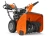 Odśnieżarka ST330 Husqvarna w kolorze pomarańczowym, ustawiona w pozycji stojącej; widok z boku ukazujący wirnik śrubowy oraz silnik. Ręczne uchwyty do prowadzenia, duże gumowe koła, na obudowie wyraźne logo Husqvarna. Tło neutralne.