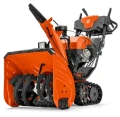 Odśnieżarka ST 427T Husqvarna w kolorze pomarańczowym z czarnymi detalami, z szeroką, zębatą strugą odśnieżającą oraz kółkami z bieżnikiem. Produkt stoi na podłożu, widok z boku. Na uchwycie znajdują się przyciski sterujące.