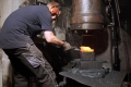 Metalowe, wąskie widełki Krumpholz do bonsai na stole w ciemnym warsztacie. Pracownik w rękawicach i ochronnych słuchawkach kształtuje metal pod prasą, z intensywnym ciepłem i iskrzeniem w tle.