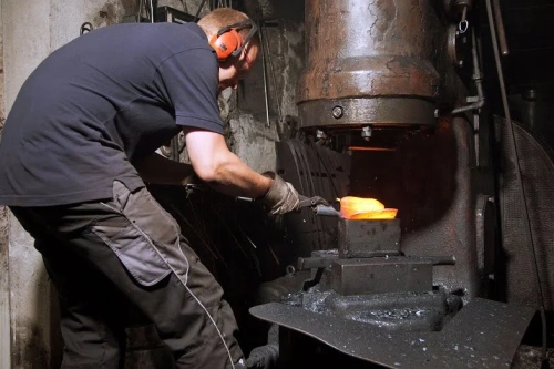 Metalowe, wąskie widełki Krumpholz do bonsai na stole w ciemnym warsztacie. Pracownik w rękawicach i ochronnych słuchawkach kształtuje metal pod prasą, z intensywnym ciepłem i iskrzeniem w tle.