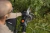 Pilarka Husqvarna Aspire™ C15X-P4A w rękach użytkownika, ujęcie z góry; narzędzie o szarym korpusie, z pomarańczowym przyciskiem uruchamiania, tnące gałęzie. Tło naturalne, trawa, a także kilka przyciętych fragmentów roślinności.