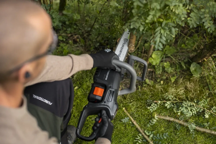 Pilarka Husqvarna Aspire™ C15X-P4A w rękach użytkownika, ujęcie z góry; narzędzie o szarym korpusie, z pomarańczowym przyciskiem uruchamiania, tnące gałęzie. Tło naturalne, trawa, a także kilka przyciętych fragmentów roślinności.
