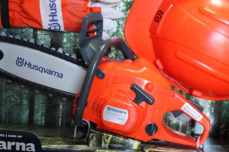 Zestaw Husqvarna dla dzieci: pomarańczowa pilarka 440, kask z logo i rękawice. Zdjęcie frontalne na tle lasu podkreśla charakter i bezpieczeństwo zabawki.